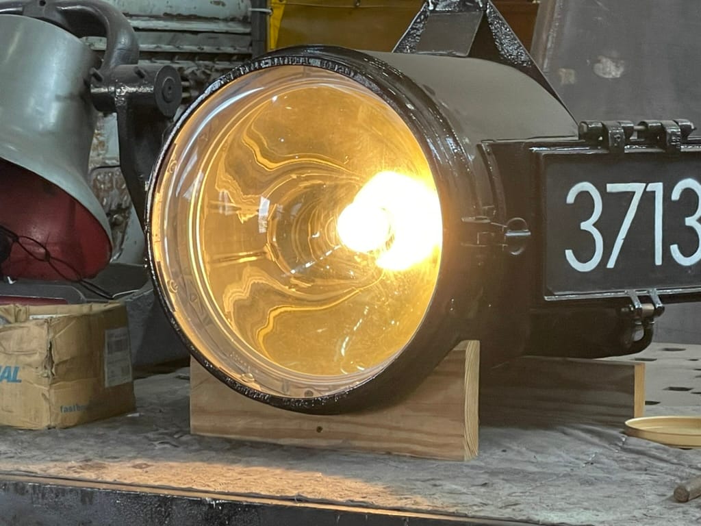 A round black headlight on a wooden stand. The light is illuminated. There is a number board that says 3713. the bell is visible on the left in the background.