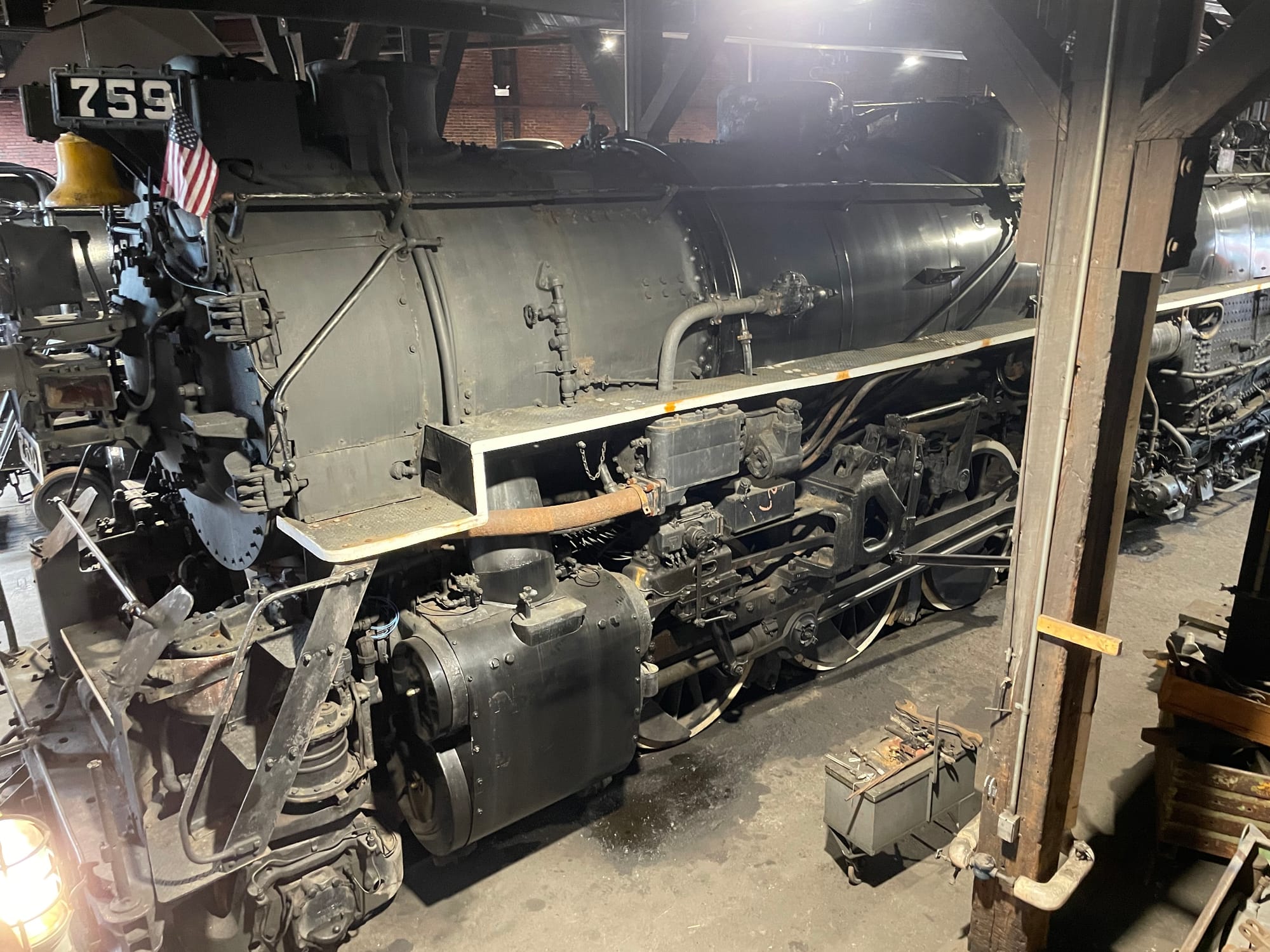 A gray and black steam locomotive in a shed. On the front of the locomotive you can see the brass bell hanging under a number board that says 759. A small American flag is next to the bell