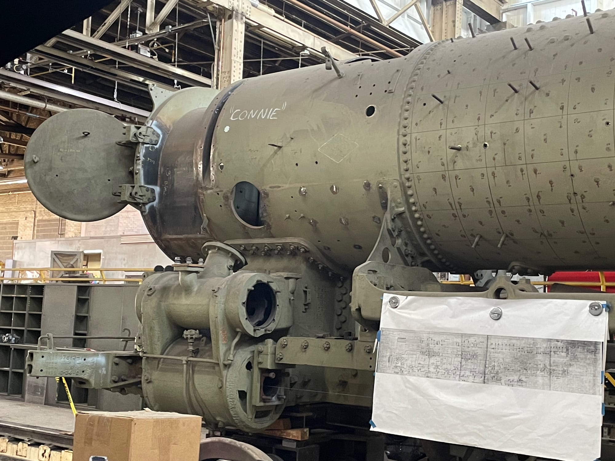 A steam locomotive under construction. The boiler has the jackets removed, exposing the gray/green skin underneath. The forward access hatch is open. The piston shapes are on the locomotive but are incomplete. Bolts and other things are protruding from the surfaces. The wheels are removed. In the foreground attached to the frame is a schematic the workers are using to put the machine back together.