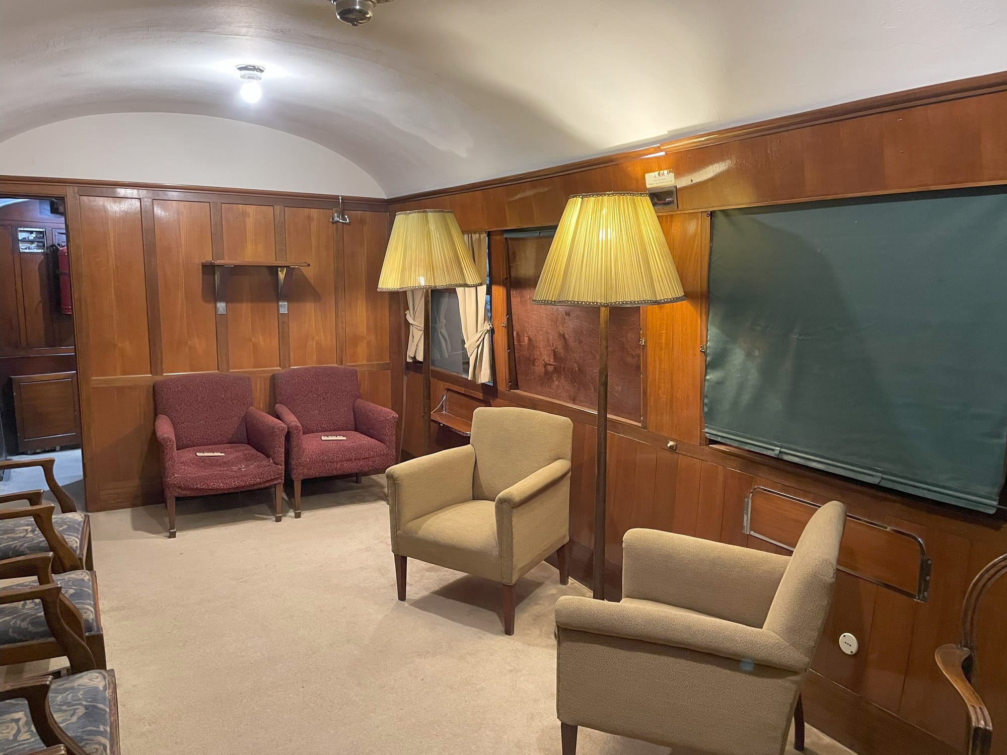 A wood paneled interior of a plush lounge car. The shades are drawn across the windows. The carpet is tan. There are two tan lounge chairs in the foreground with a lamp between them. There's two red lounge chairs in the background with another lamp.