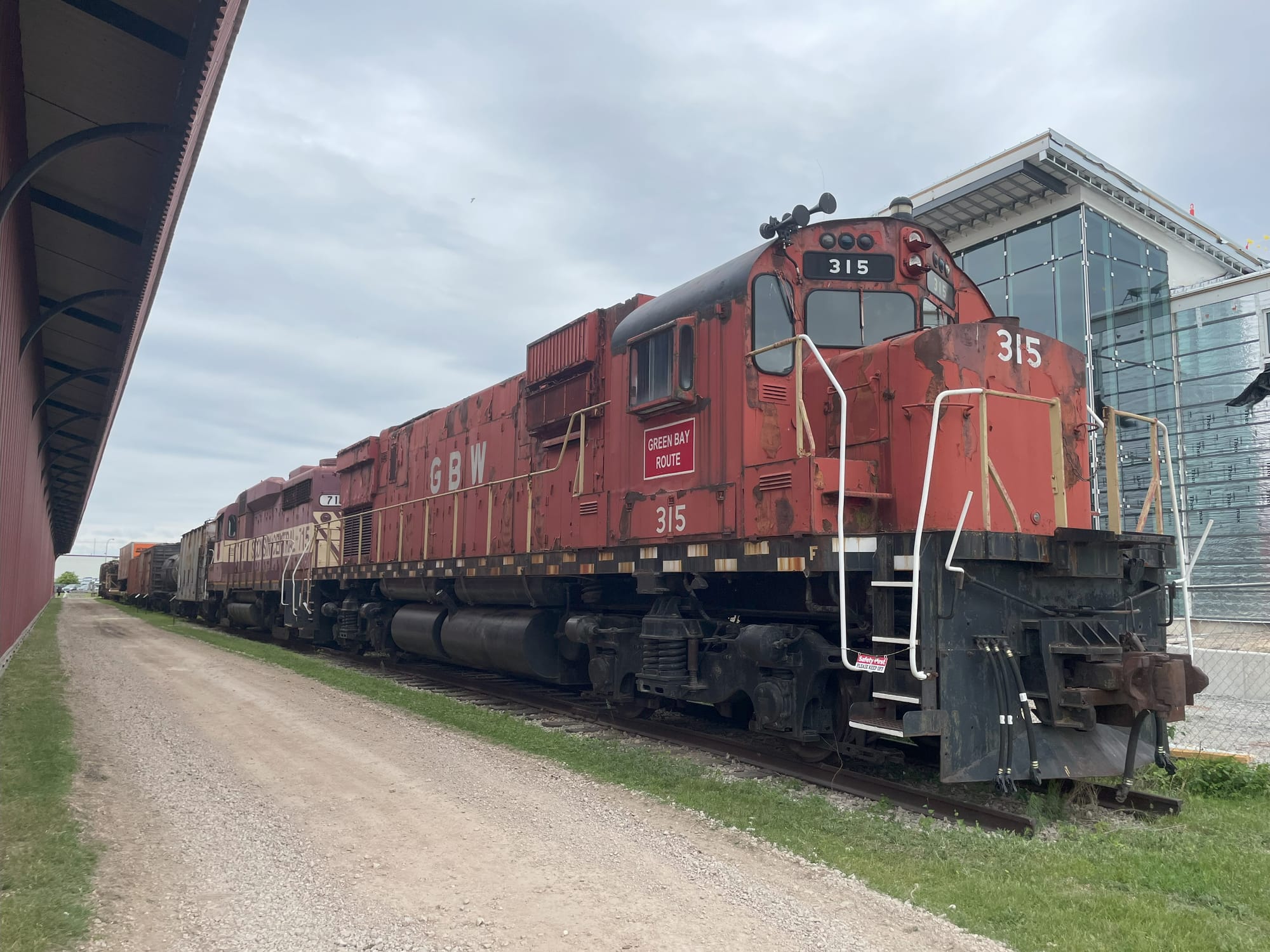 A red diesel locomotive "Green Bay Round 315" in front of a glass building under construction