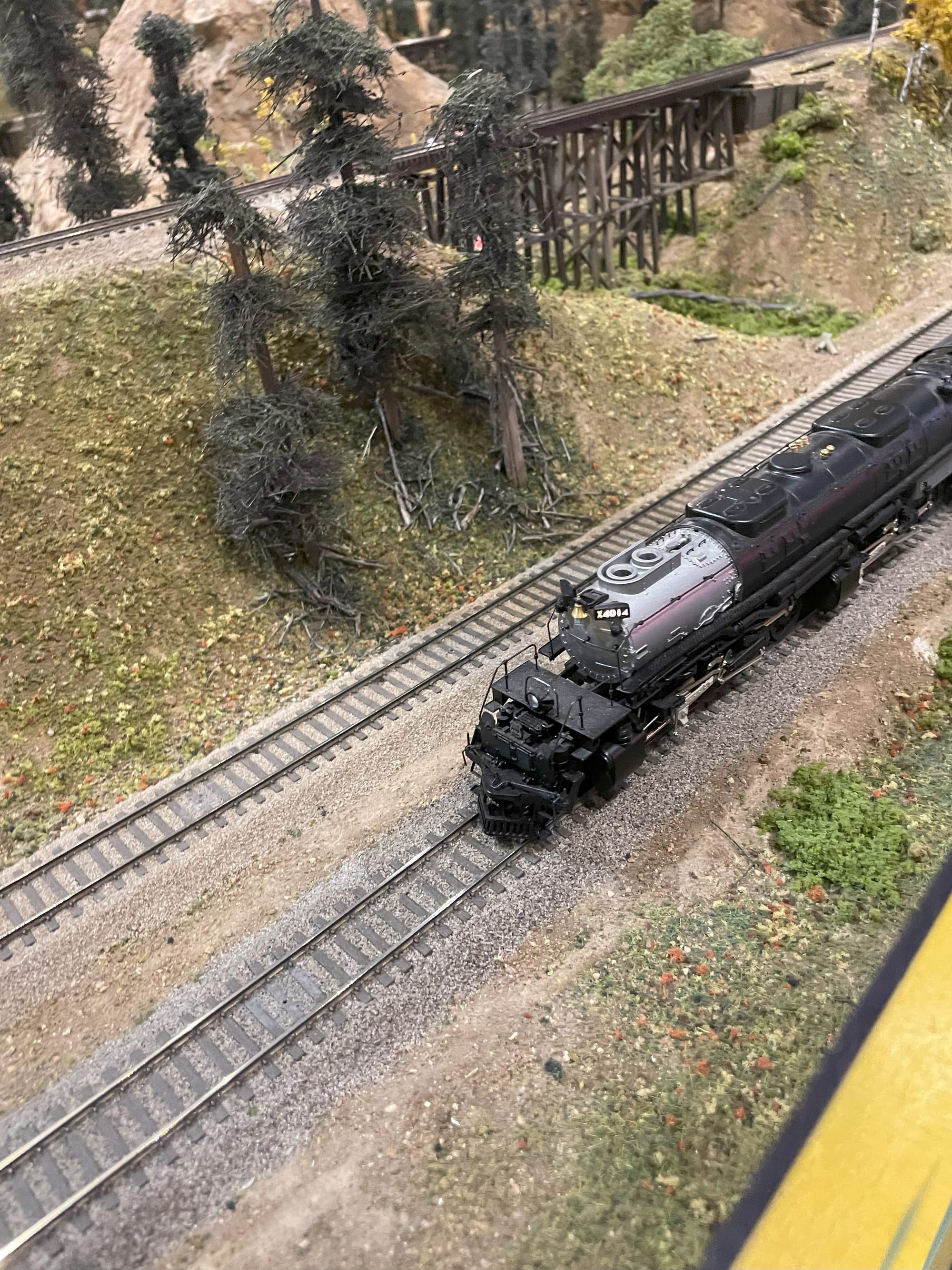 A model of Big Boy #4014 pictured from above. You can see the silver and black boiler rendered in plastic on an HO size track. There are trees and a wooden bridge in miniture in the background