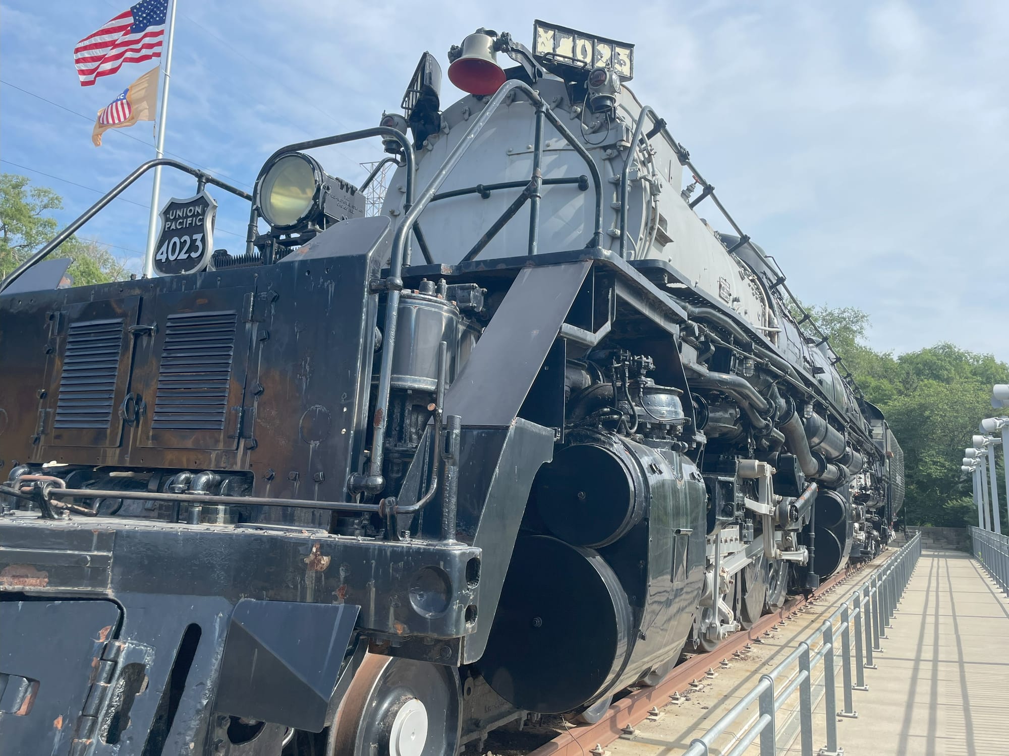 A steam locomotive with a black frame and silver boiler. The shield on the front says Union Pacific 4023 below a headlight. In the background an American Flag flies above a yellow flag wih the red-white-blue logo of Union Pacific