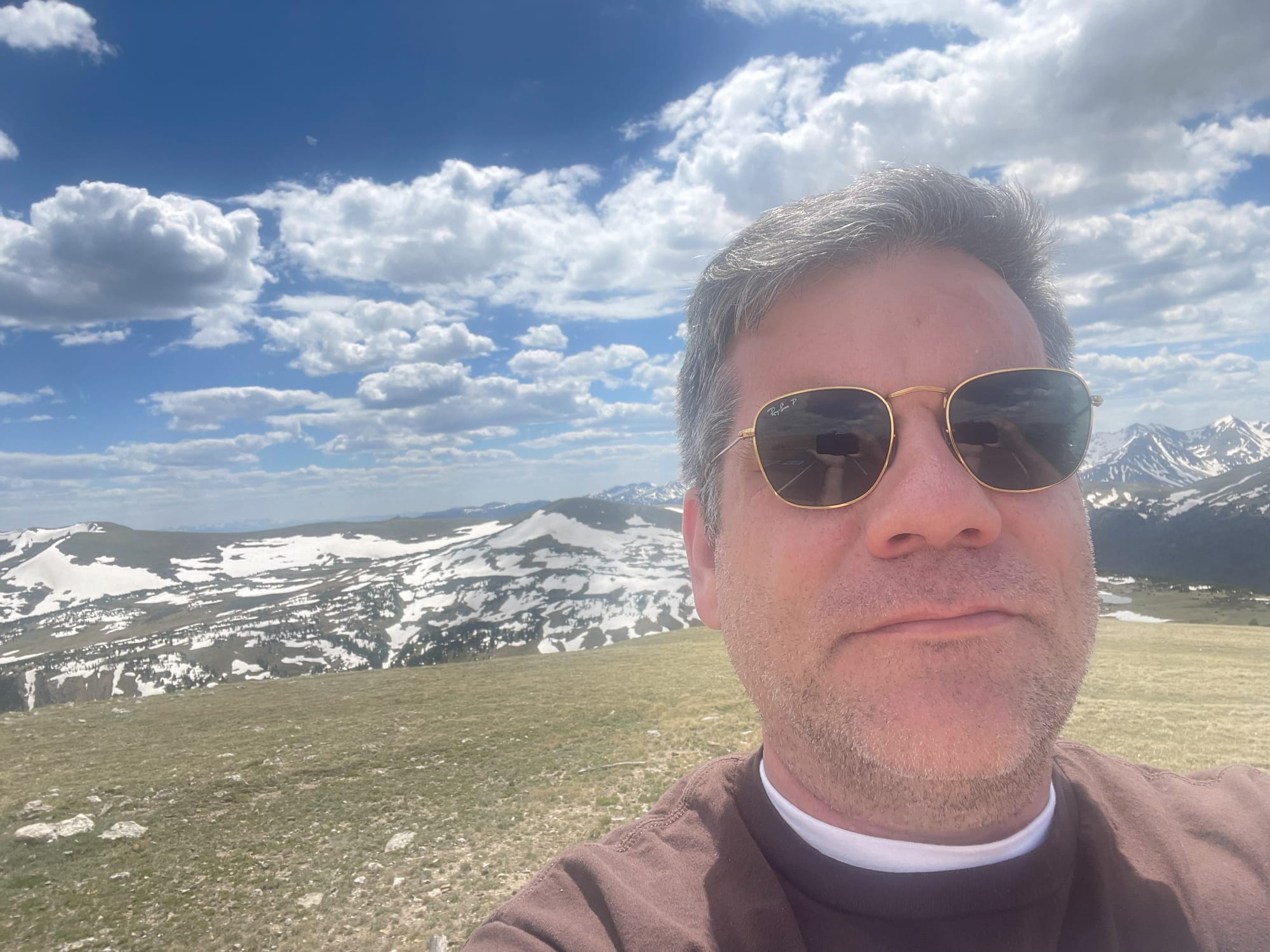 A middle-aged man taking a selfie like he's a kid. He's got sunglasses on, somewhat silver hair, and gray stubble. He's wearing a brown shirt. In the background is snowcapped mountains