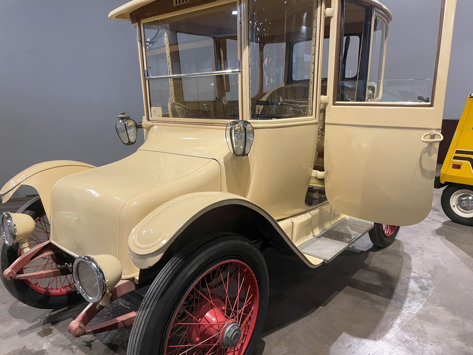  A cream colored antique car. The hood is short while the cabin area has a vertical windshield. The doors are similar to stage coach doors (like on the Model A). The wheels are spoked and painted red. It has headlights separate from the body and mounted low in front of the fenders. In place of side mirrors like on a modern car, it has lanterns