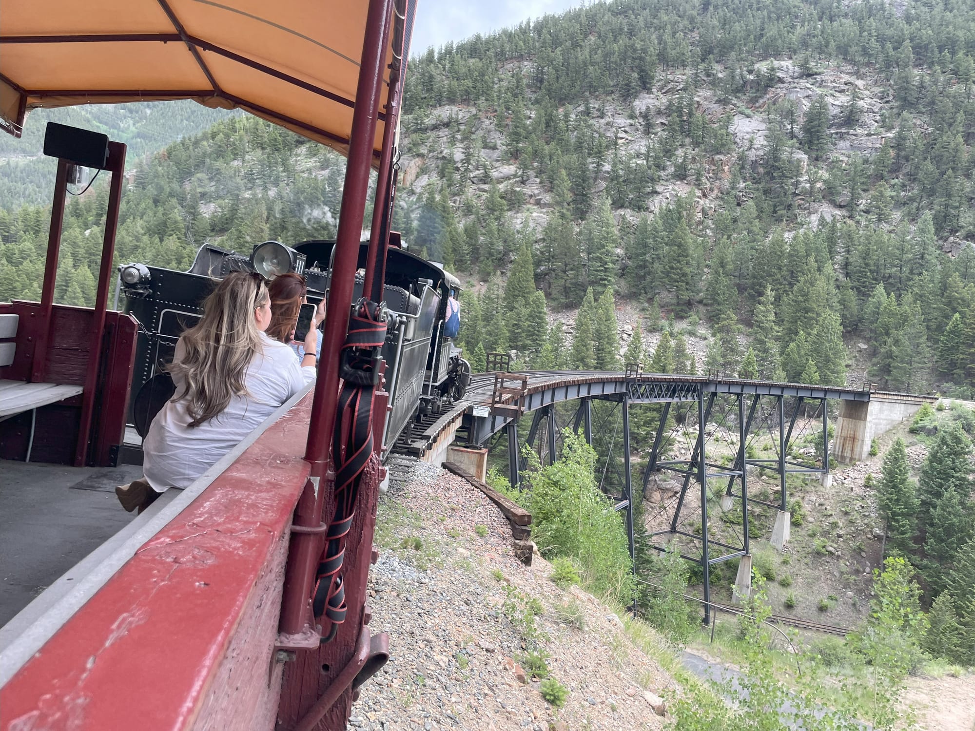 A view from the first car behind a steam locomotive going over a bridge. Tracks the train had run on are visible under the bridge, with the pine trees of a mountain in the background.