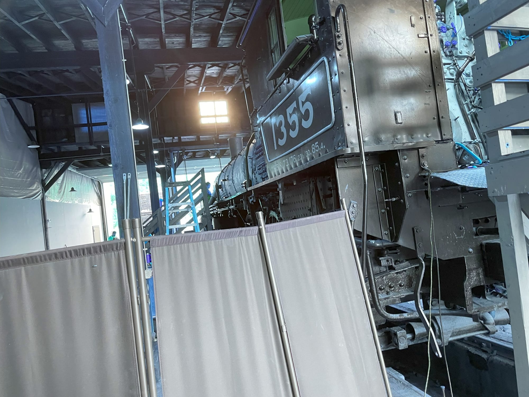 A steam locomotive cab with the #1355 printed on the side. The boiler piece in the background is exposed. Beyond the locomotive the sun is streaming through a window near the ceiling. A canvas screen in the foreground obscures the locomotive's wheels