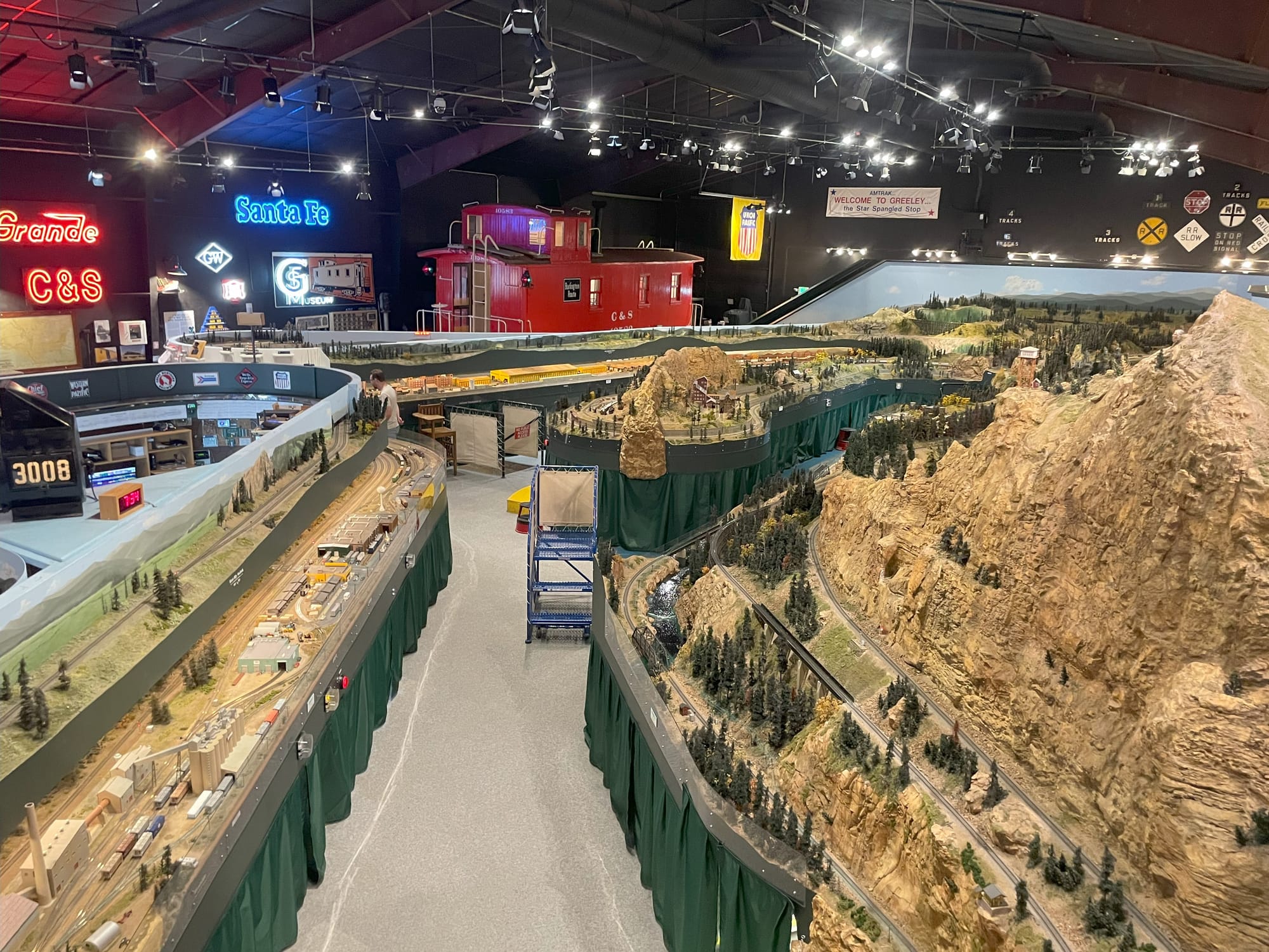 A giant train layout from a bove. The picture shows two mountain areas with tracks wrapping aroudn them. In the background is a red caboose. Several lighted signs for various railroads including C&S, Rio Grande, and Santa Fe are on the walls