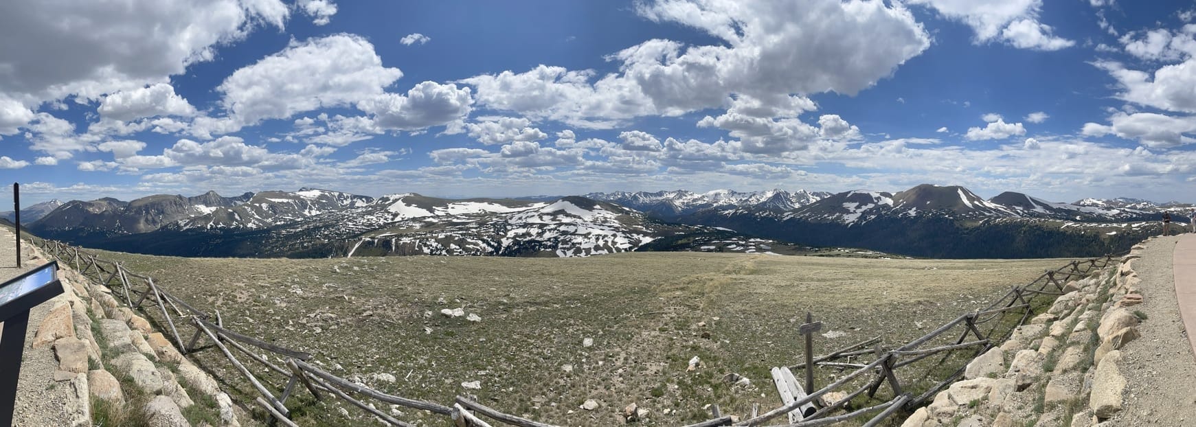 A panorama of mountains. In the foreground a wooden fence in front of some tundra beore a drop off and snowcapped mountains in the background under a blue sky with puffy clouds