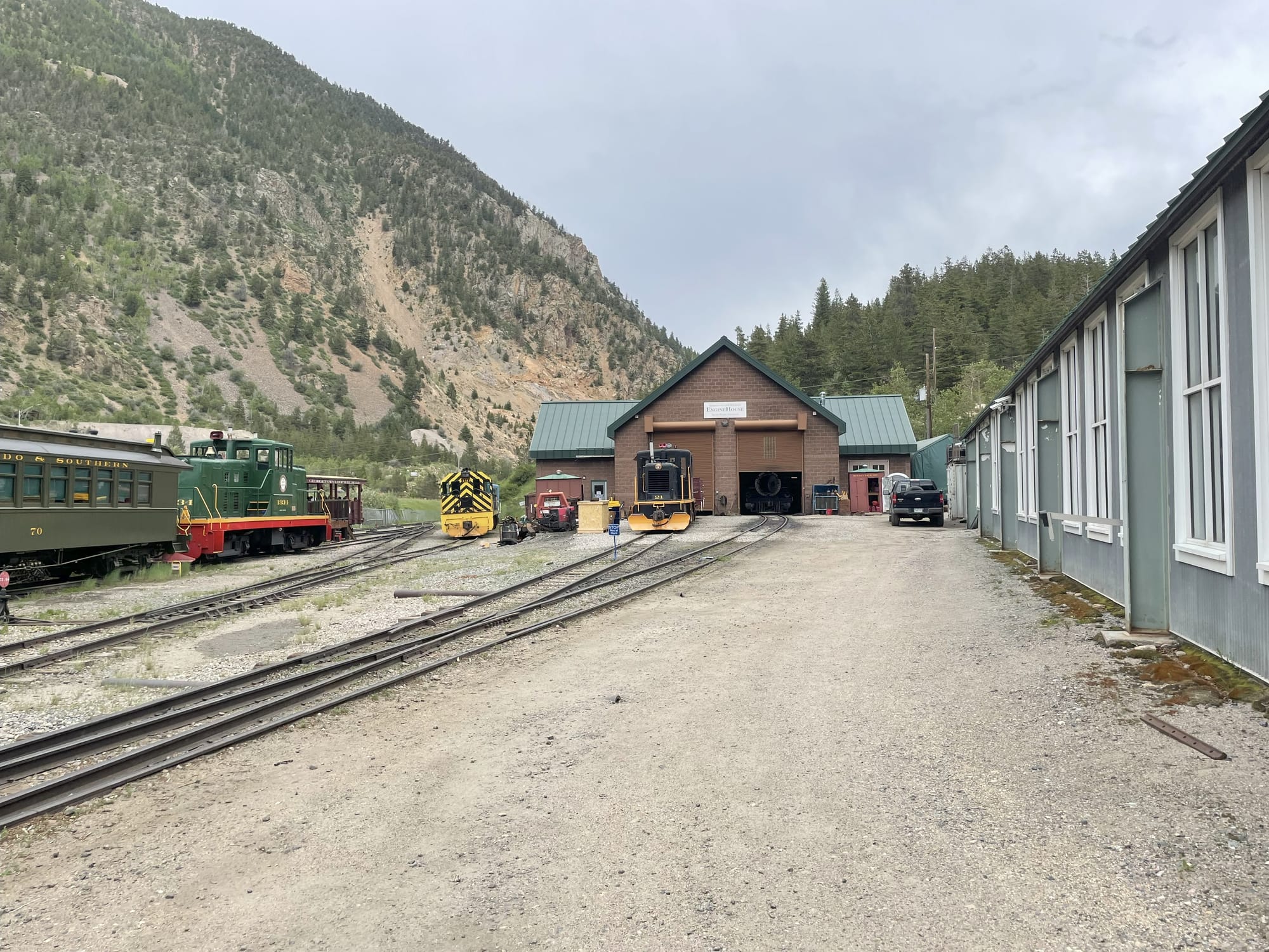 A small railroad yard in the mountains. A building with tracks going into it is in the background. There's several diesel switching locomotives. One black and yellow. One black with yellow stripes. One green and red pulling a green passenger car. The engine shed is red brick with a green roof