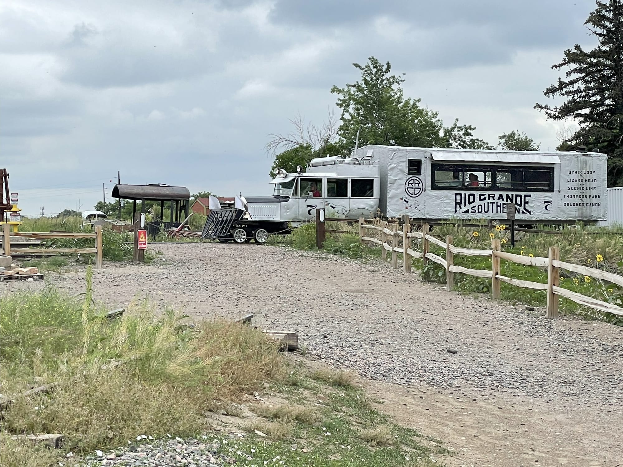 A silver railcar with an old road car construction on the front with a black cow catcher. The cab looks like a semi truck and the backend is a box with a window. People are sitting inside. 