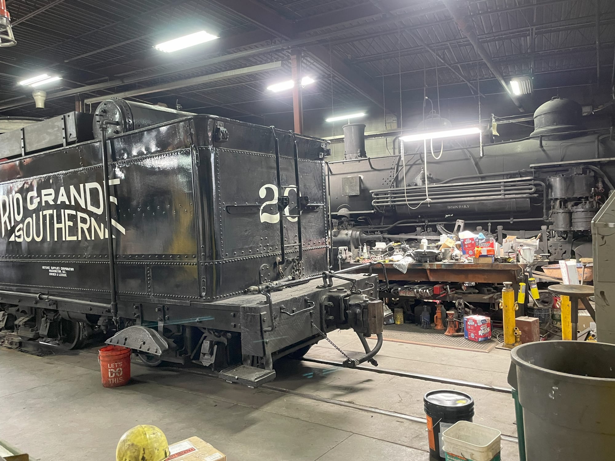A locomotive shop. In the foreground is a tender on rails built into the floor. It has a logo on the side "Rio Grande Southern" in a stylized curve pattern that looks like a hill or mountain. The number 20 is printed on the backend behind a latter. In the background the locomotive itself is visible behind a table full of tools and materials