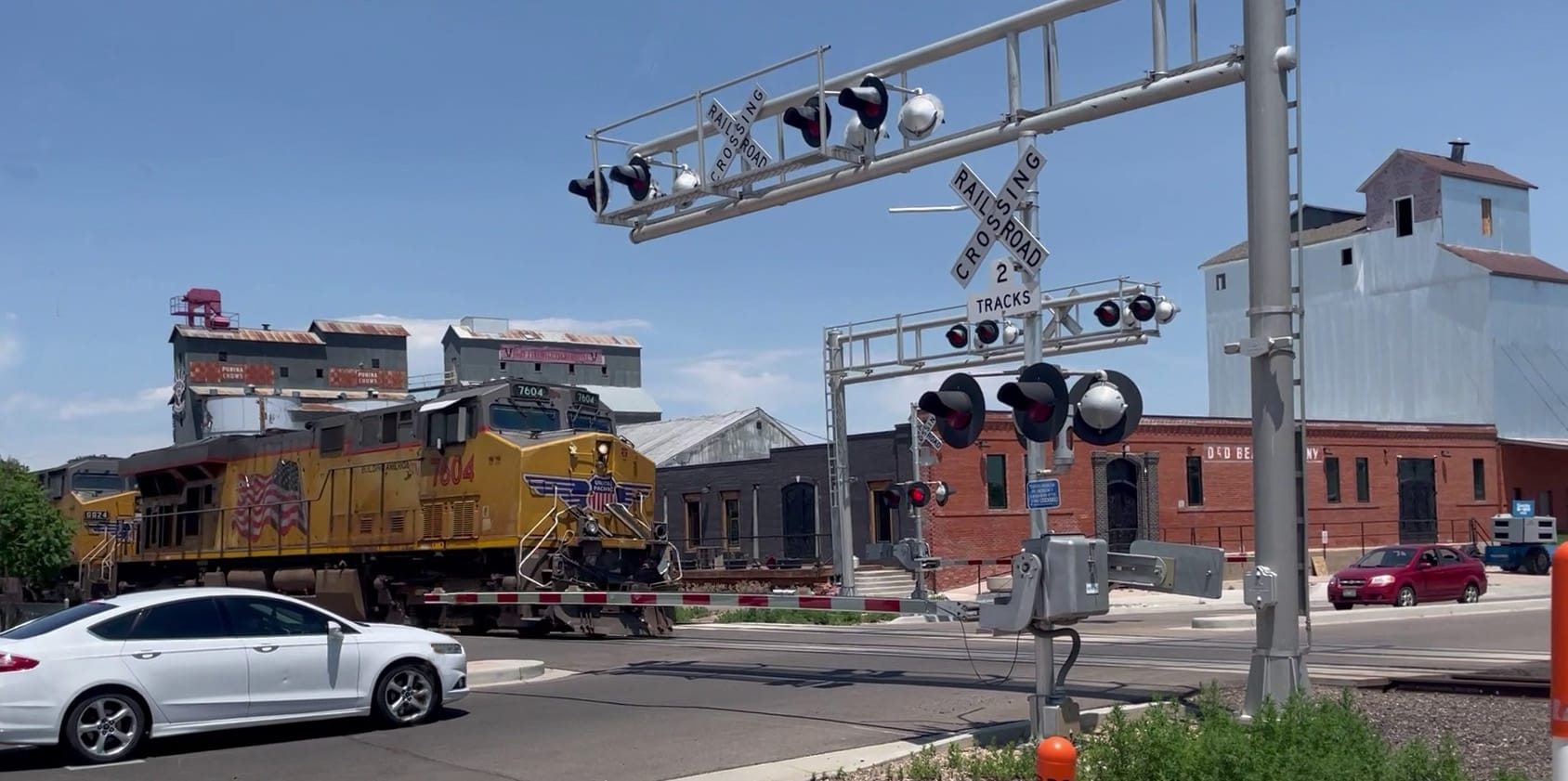 A yellow and gray diesel locomotive with a white car in the foreground. There's a automated railroad crossing with flashing lights and a red and white gate across the road. In the background there are brick and metal industrial buildings