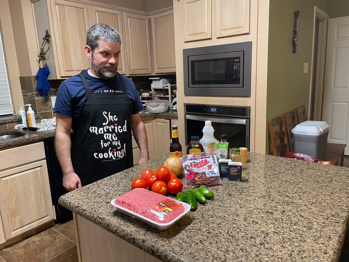 A man looking over peppers, tomatoes, spices and and meat for chili. His apron reads "She married me for my cooking".