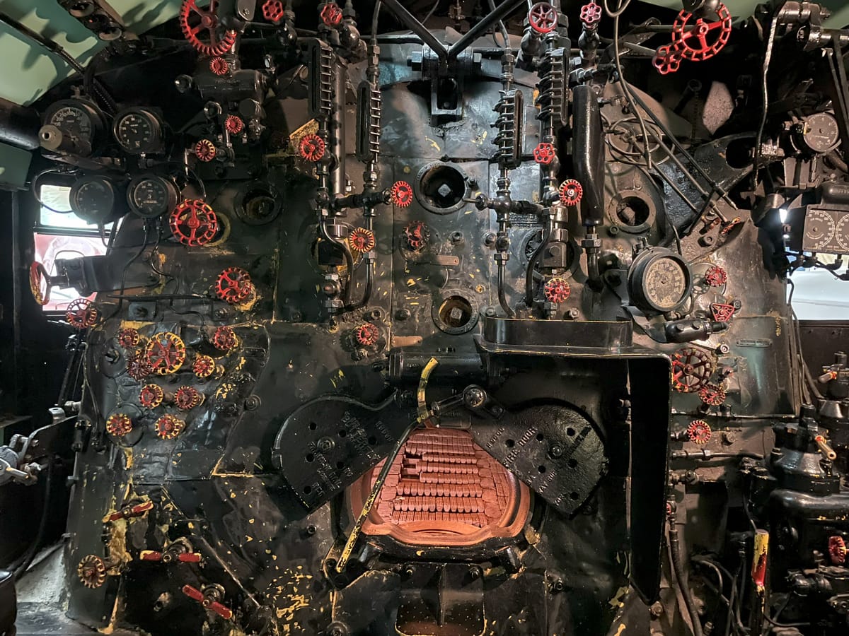 A steam locomotive cab. At the bottom center the firebox doors are open and you can see the bricks inside. Attached to the rest of the gray boiler structure has various sizes of red faucets